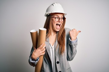 Young beautiful redhead architect woman wearing glasses and safety helmet holding blueprints pointing and showing with thumb up to the side with happy face smiling