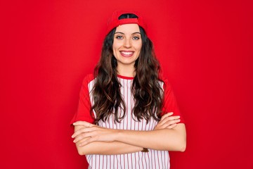 Beautiful woman with blue eyes wearing baseball sportswear and cap over red background happy face smiling with crossed arms looking at the camera. Positive person.