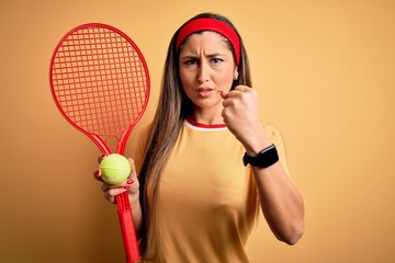 Beautiful brunette sportswoman playing tennis using racket and ball over yellow background annoyed and frustrated shouting with anger, crazy and yelling with raised hand, anger concept