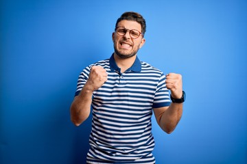 Young man with blue eyes wearing glasses and casual striped t-shirt over blue background excited for success with arms raised and eyes closed celebrating victory smiling. Winner concept.