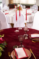 Place at the banquet table: the menu card lies on a red napkin on plates, a gilt fork and knife on the sides of the plate, a glass of brown glass, flower arrangement, red candles