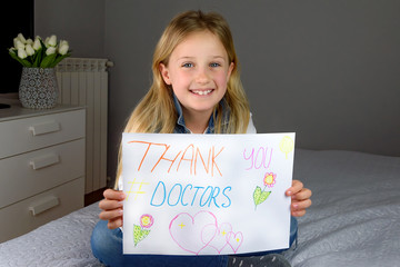 Little smiling blond girl of 9 years old sits on the bed and holds "Thank you, doctors" poster. The concept of gratitude for the fight against the virus and diseases