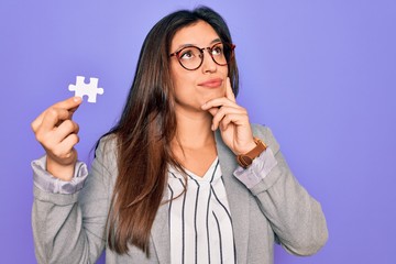 Young hispanic business woman holding puzzle peace as cooperation and union on work serious face thinking about question, very confused idea