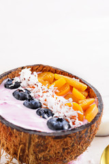 vertical smoothie bowl with frozen banana, blueberries, physalis and coconut shavings in coconut bowl with spoon on white background