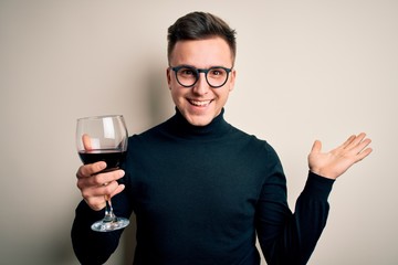Young handsome caucasian man drinking an alcoholic glass of red wine over isolated background very happy and excited, winner expression celebrating victory screaming with big smile and raised hands