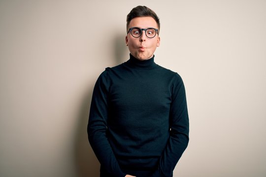 Young handsome caucasian man wearing glasses and casual sweater over isolated background making fish face with lips, crazy and comical gesture. Funny expression.