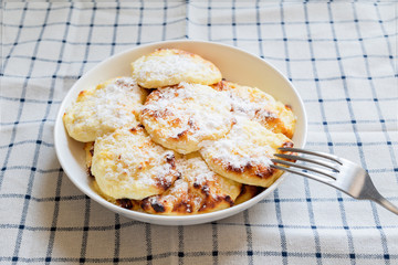 Simple breakfast for children. Sweet cottage cheese pancakes on a white plate, sprinkled with powdered sugar. Syrniki, curd fritters, baked in the oven on a checkered tablecloth background. 