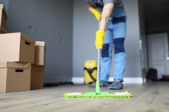 Close-up Of Professional Cleaner Washing Floor With Green Mop. Man In Working Uniform. Handyman In Gloves And Cardboard Boxes. Cleaning Service And Moving Day Concept