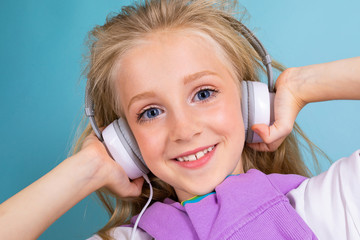 Naklejka premium portrait of a beautiful blond charming young cute sweet smiling child in a casual look with white headphones listening to the music and on a light blue background
