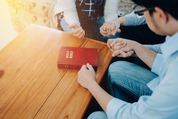 Christian small group holding hands and praying together around a wooden table with the bible on...