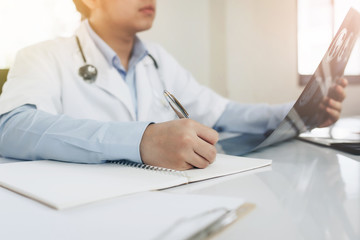 Young Asian doctor with Medical stethoscope holding X-ray film while writing on notebook  in an office at the hospital