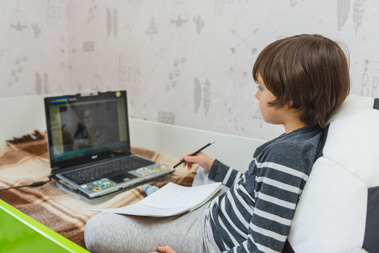 Distance Learning For Children During The Coronavirus Epidemic. The Boy Sits On The Bed And Receives A School Assignment Using The Internet And A Laptop.