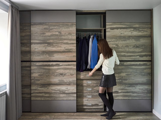 Stylish young woman standing with her back looks into an open wardrobe with clothes and opens...