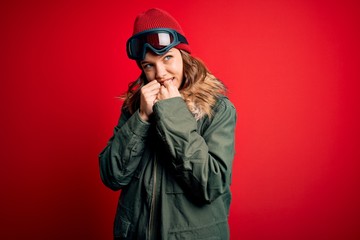 Young blonde girl wearing ski glasses and winter coat for ski weather over red background laughing nervous and excited with hands on chin looking to the side