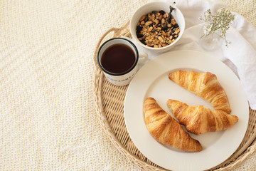 Close up of tray with coffee cup, croissants, muesli and white flowers on knitted blanket at cozy scandy boho bedroom. Bed breakfast, slow living, jomo, morning, holidays concept. Copy space, top view