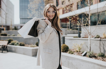 Attractive cute girl with the shopping bags and warm drink in her hand. Modern city megapolis on background. Lady order and shop online. Beautiful caucasian lady