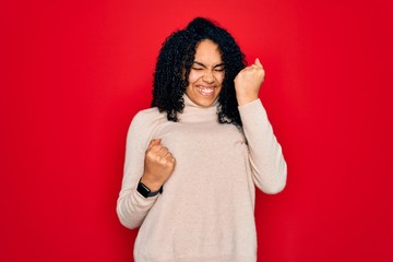 Young african american curly woman wearing casual turtleneck sweater over red background celebrating surprised and amazed for success with arms raised and eyes closed. Winner concept.