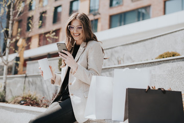 Attractive cute girl with the shopping bags and smarphone in her hand. Modern city megapolis on background. Lady making a phone call to order and shop online. Beautiful caucasian lady