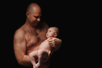 father holds a newborn son in his arms. Bald man with a naked torso and a 4 month old baby on a black background