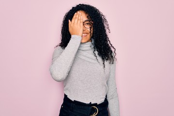 Young african american woman wearing turtleneck sweater and glasses over pink background covering one eye with hand, confident smile on face and surprise emotion.