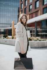 Attractive cute girl with the shopping bags and warm drink in her hand. Modern city megapolis on background. Lady order and shop online. Beautiful caucasian lady