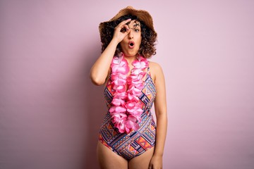 Young beautiful arab woman on vacation wearing swimsuit and hawaiian lei flowers doing ok gesture shocked with surprised face, eye looking through fingers. Unbelieving expression.