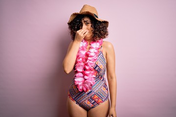 Young beautiful arab woman on vacation wearing swimsuit and hawaiian lei flowers smelling something stinky and disgusting, intolerable smell, holding breath with fingers on nose. Bad smell