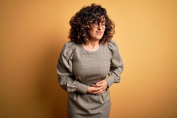Beautiful arab business woman wearing dress and glasses standing over yellow background with hand on stomach because indigestion, painful illness feeling unwell. Ache concept.