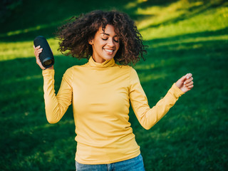 Modern trendy african american girl listening to music by wireless portable speaker.Young beautiful woman enjoying, dancing in park.