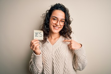 Young beautiful woman with curly hair holding reminder paper with yes word message with surprise...