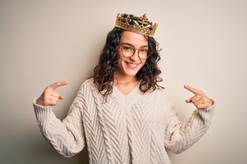 Young beautiful woman with curly hair wearing golden queen crown over white background looking...