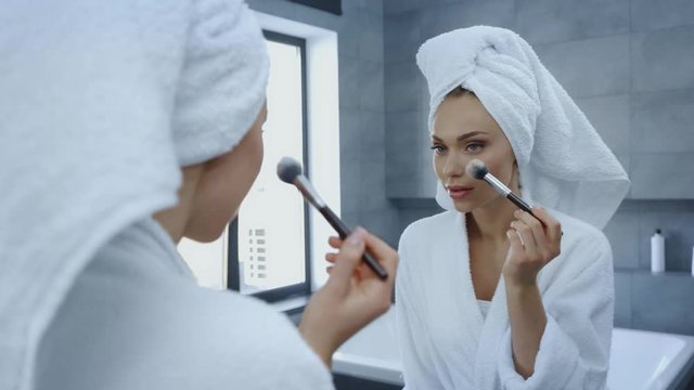 beautiful young woman in bathrobe using cosmetic brush in front of mirror