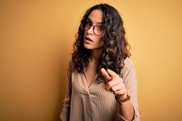 Fototapeta premium Beautiful woman with curly hair wearing striped shirt and glasses over yellow background pointing displeased and frustrated to the camera, angry and furious with you