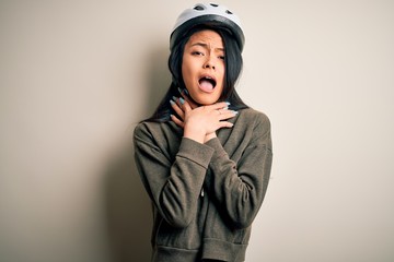 Young beautiful chinese woman wearing bike helmet over isolated white background shouting and suffocate because painful strangle. Health problem. Asphyxiate and suicide concept.