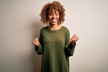 Beautiful african american woman with curly hair wearing casual sweater over white background celebrating surprised and amazed for success with arms raised and eyes closed. Winner concept.