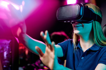 Positive girl using virtual reality simulator gesturing hands to bright purple light while playing...