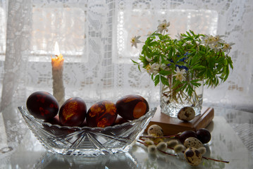 A bouquet of spring flowers in a glass, painted Easter eggs on a knitted napkin against a background of white lace curtains.