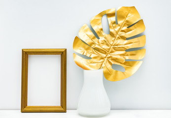 gold and white interior mockup design. Empty photoframe and monstera leaf im white vase on white wall background. Trendy design