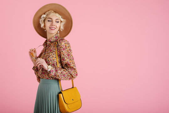 Fashionable happy smiling woman wearing trendy spring, summer outfit: hat, floral print blouse, pleated skirt, with yellow shoulder bag, posing on pink background. Copy, empty space for text