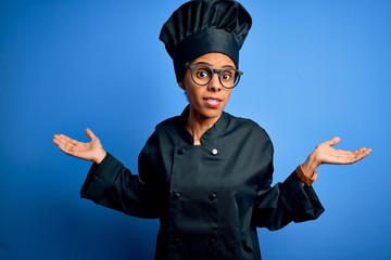 Young african american chef woman wearing cooker uniform and hat over blue background clueless and confused expression with arms and hands raised. Doubt concept.