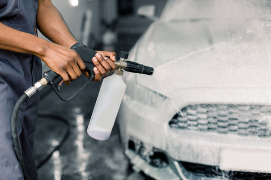 Manual car washing concept. Cropped image of hands of young dark skinned man worker cleaning modern white car with foam and pressured water at service station. Manual detailing car wash