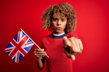 Young african american curly woman holding uk flag celebrating brexit referendum pointing with finger to the camera and to you, hand sign, positive and confident gesture from the front