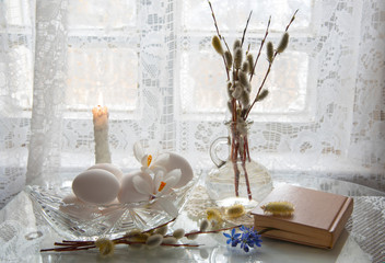White eggs in a crystal vase, willow branches in a bottle and an old book on a light background.