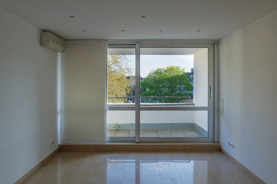 Interior view of empty room without people, with air condition, white wall, tile floor and aluminium sliding windows and background of treetop.