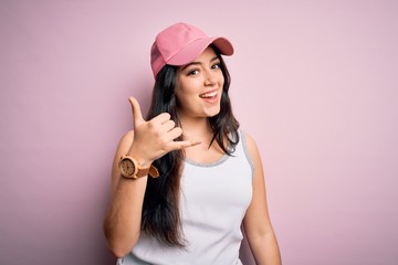 Young brunette woman wearing casual sport cap over pink background smiling doing phone gesture with hand and fingers like talking on the telephone. Communicating concepts.