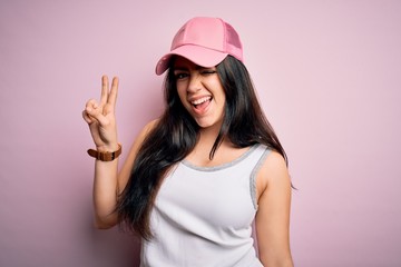 Young brunette woman wearing casual sport cap over pink background smiling with happy face winking at the camera doing victory sign. Number two.