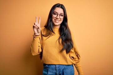 Young brunette woman wearing glasses and casual sweater over yellow isolated background smiling looking to the camera showing fingers doing victory sign. Number two.