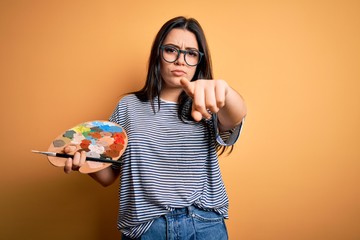 Young brunette artist woman painting holding painter brush and palette over yellow background pointing with finger to the camera and to you, hand sign, positive and confident gesture from the front