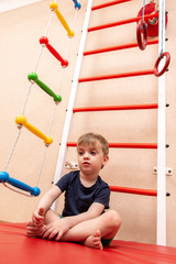 little boy sits on a red mat near the Swedish wall. Doing sports at home