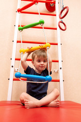 a three-year-old child sits on a sports mat near the Swedish wall